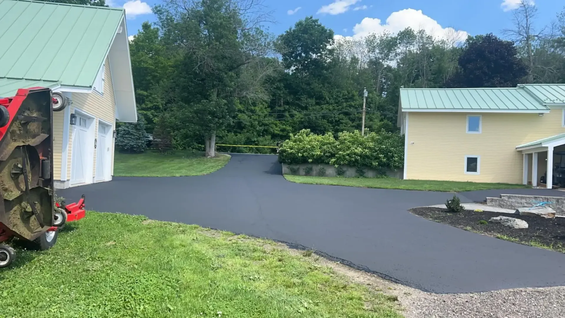 Newly paved asphalt driveway, with two buildings in the background, and landscaping to the right.