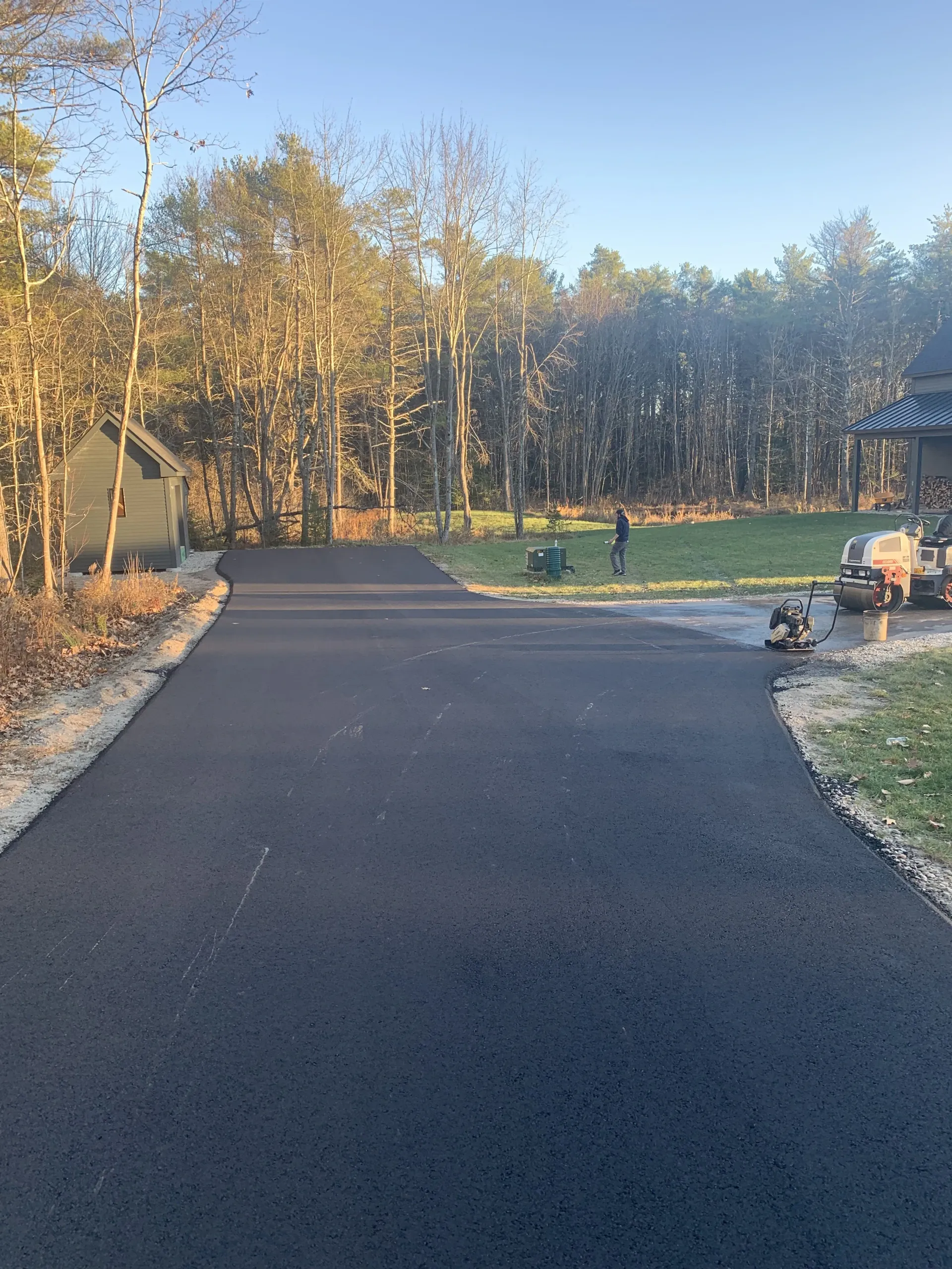 Freshly paved asphalt driveway leads to a yard with trees; a shed and workers are visible in the distance.