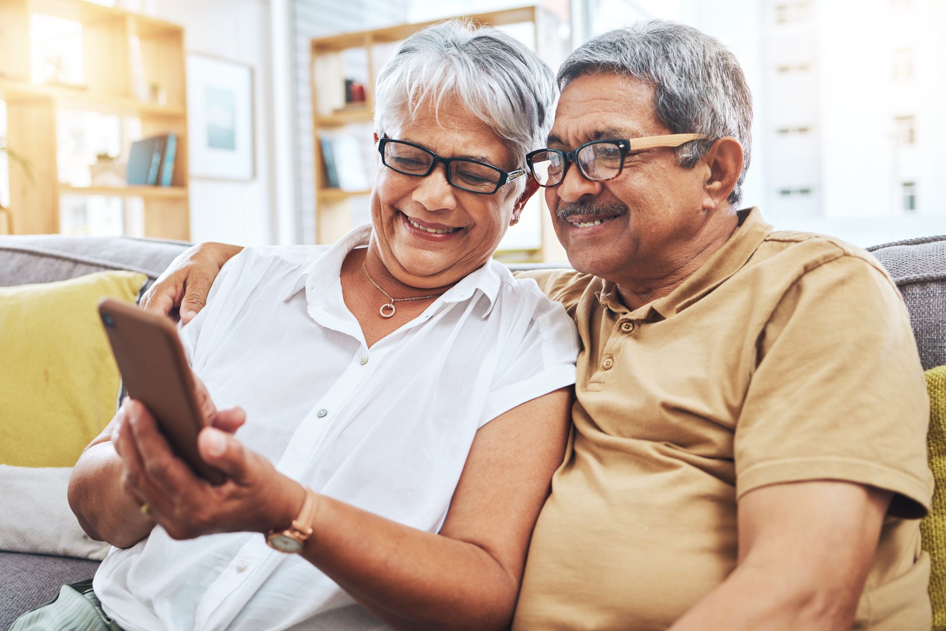 Two smiling people sit on a couch, looking at a smartphone together in a bright living room.