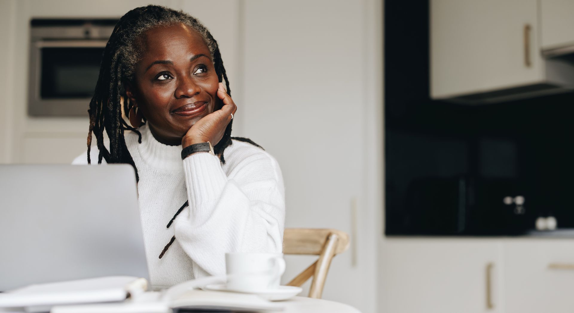 Woman smiling at a laptop in a bright kitchen, resting her chin on her hand.