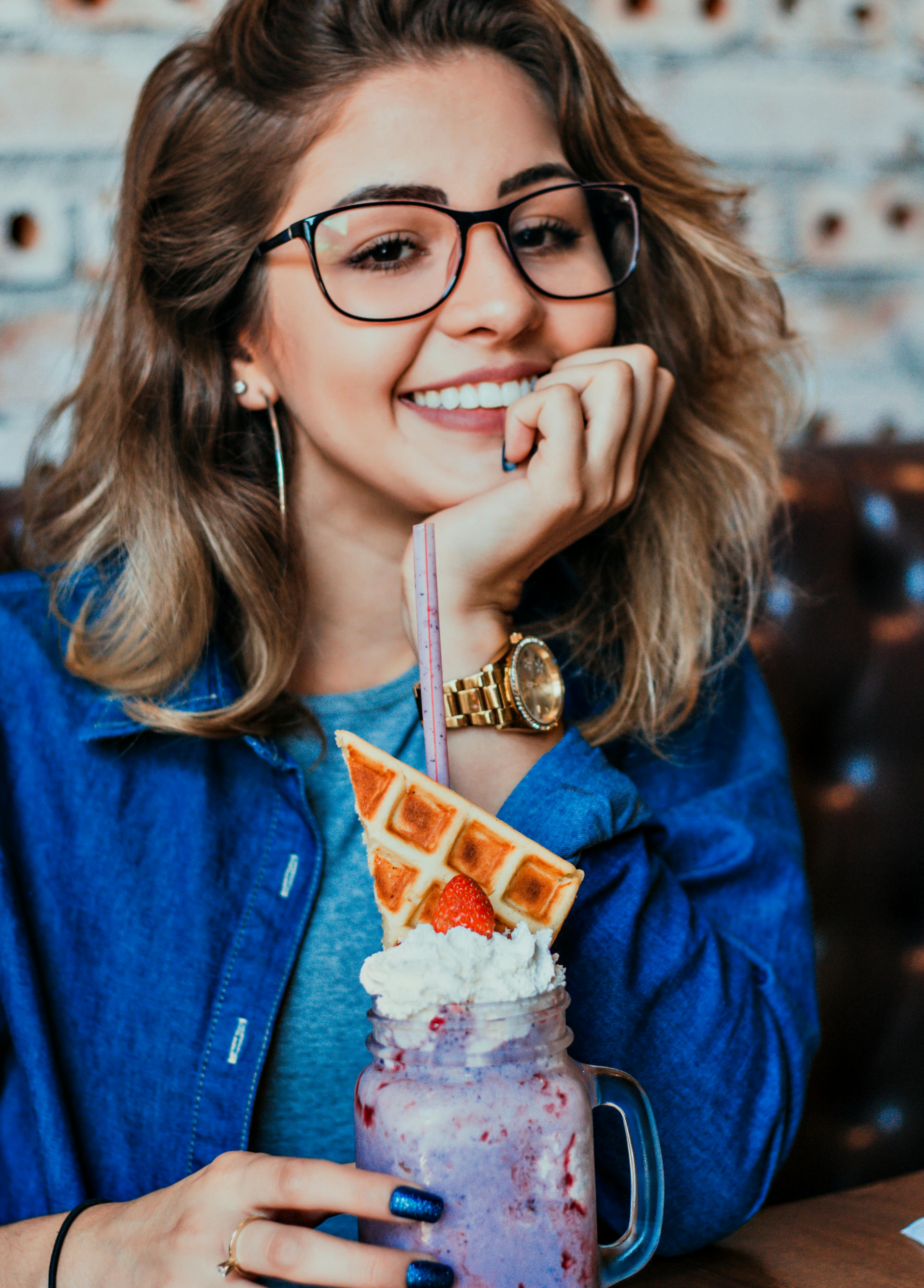 Smiling person in glasses holding a tall purple drink topped with whipped cream and waffle cones