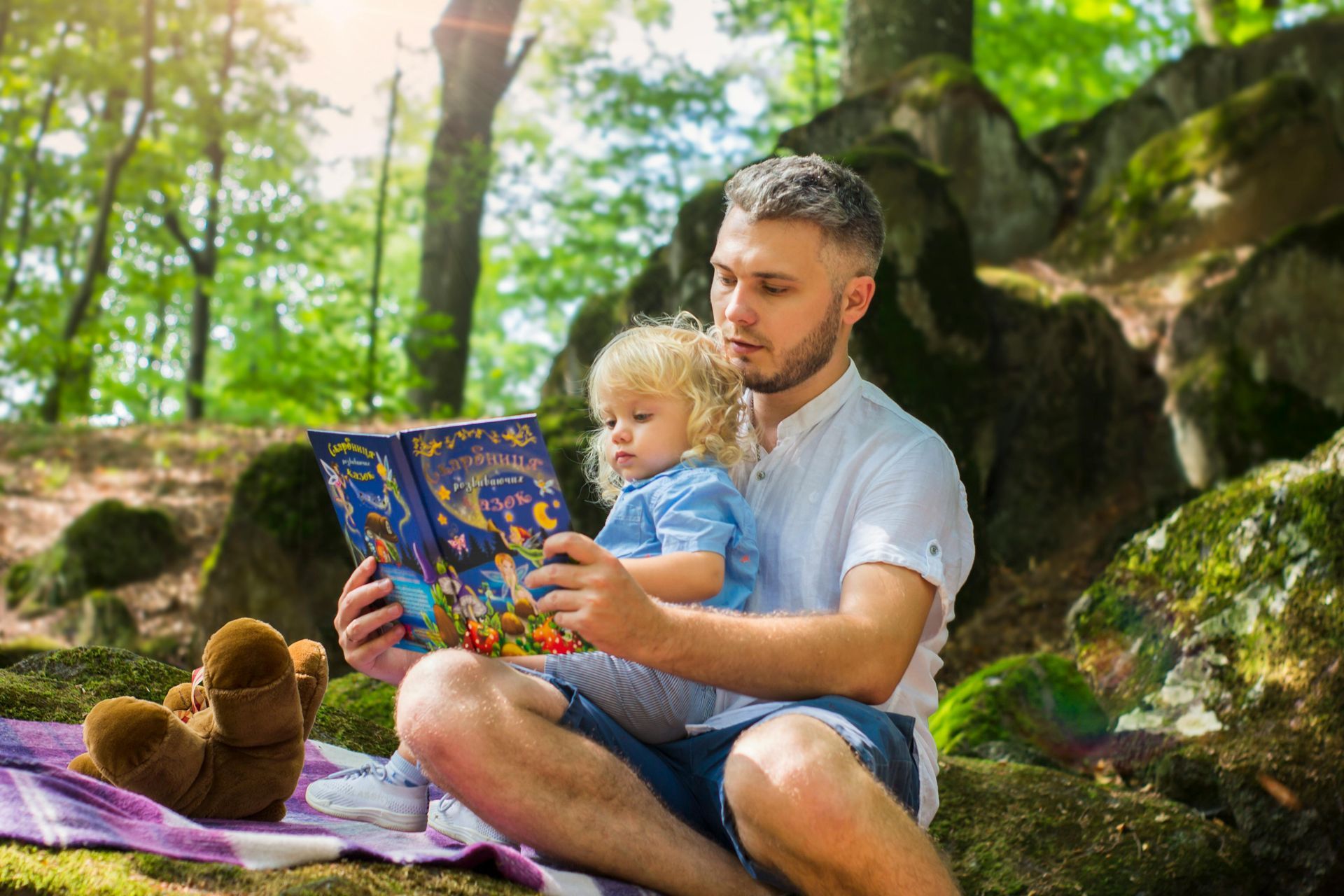 Man reading a colorful book to a child on a blanket in a leafy park