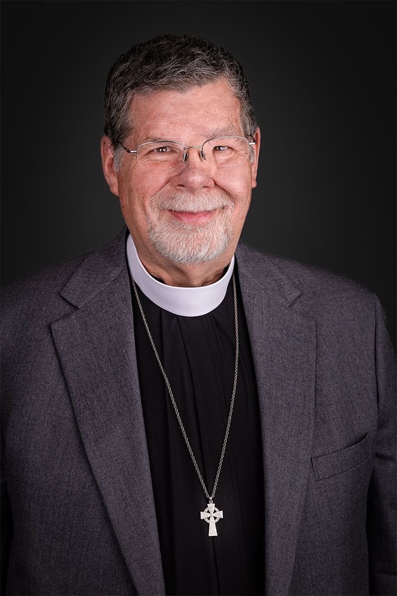 Man wearing a clerical collar, eyeglasses, and blazer, smiling.