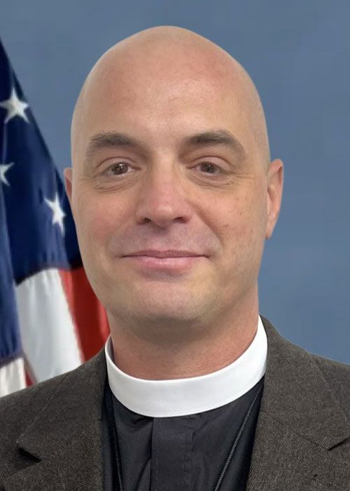 Bald man in clerical collar and jacket, US flag in background.