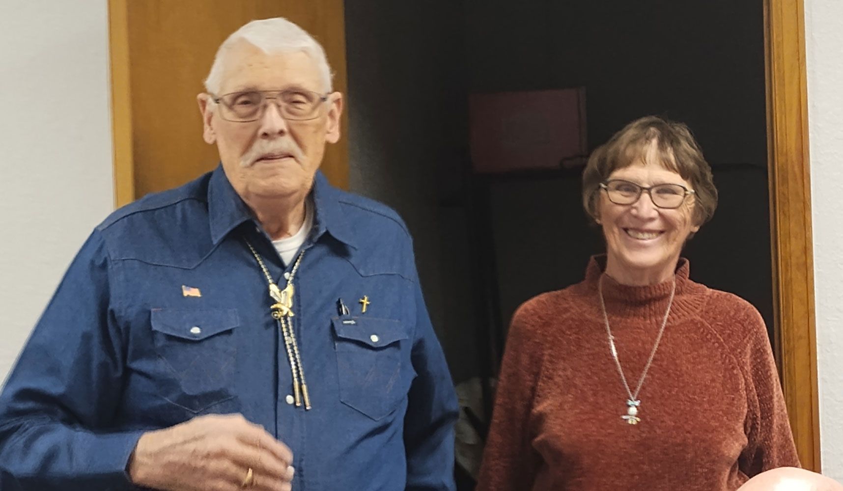 An older man in a blue shirt with a bolo tie and an older woman in a brown sweater smile side-by-side indoors.