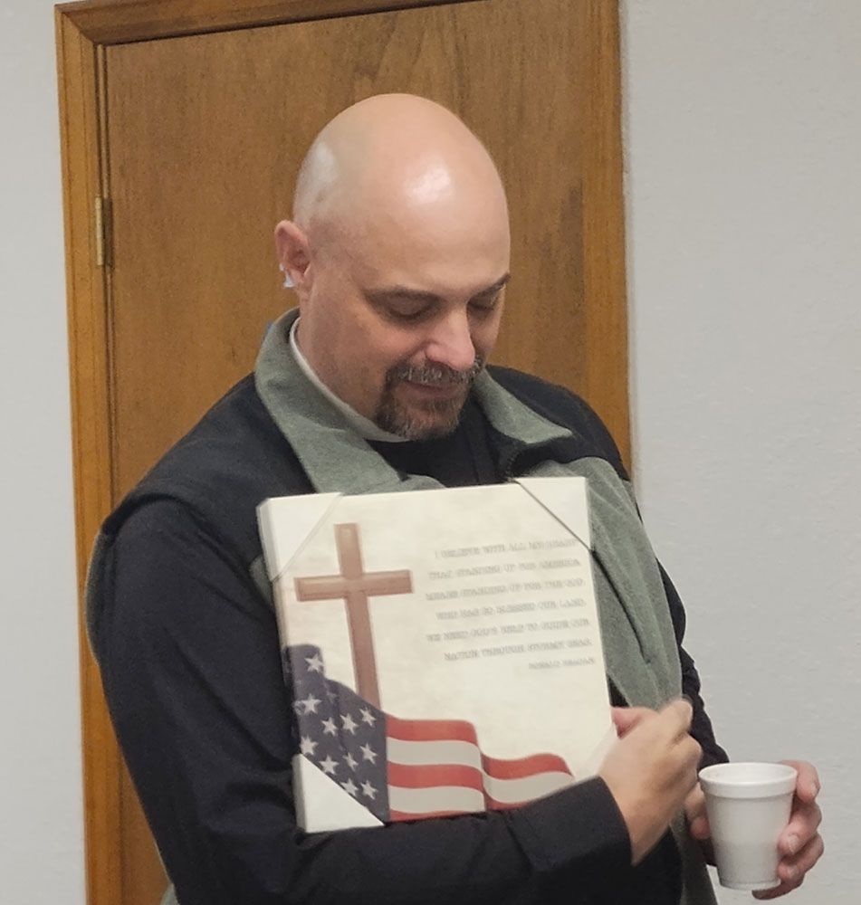A person holding a square canvas featuring a cross and an American flag with text, while holding a white disposable cup.