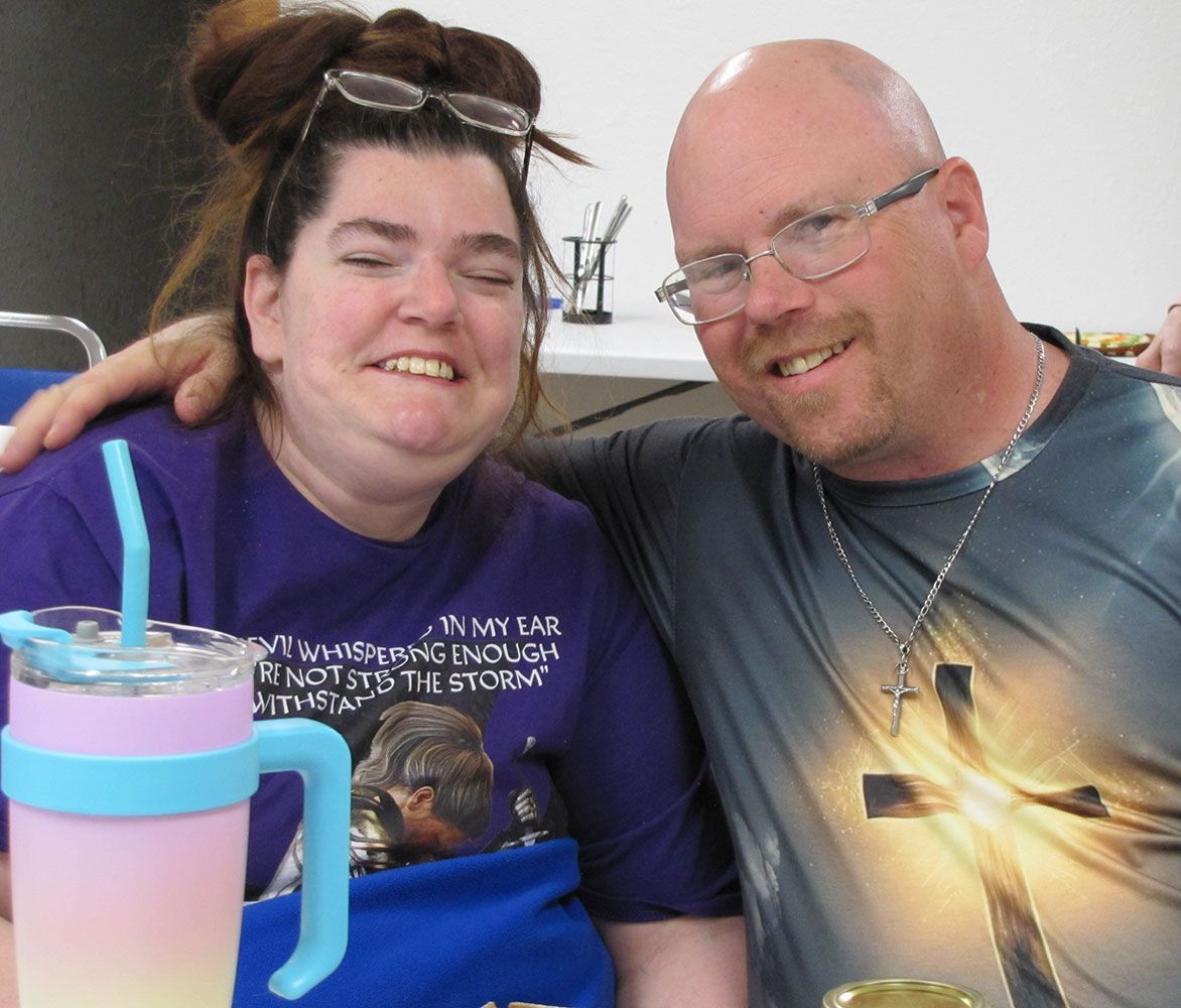 A smiling person with glasses and a purple shirt sits next to a person with glasses and a cross-print shirt; arm around.
