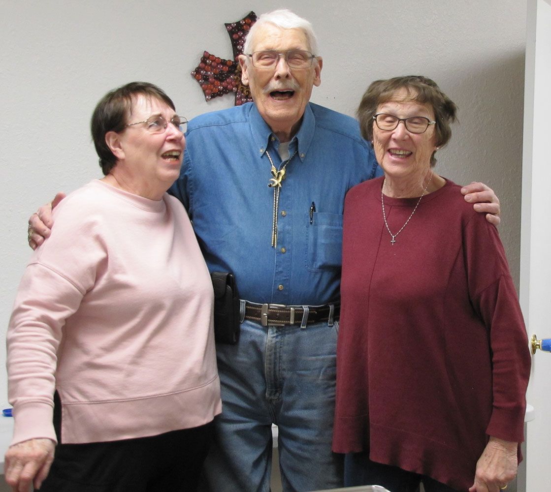 Three people stand close together smiling, with the central person in a blue shirt and the two others in pink and maroon.