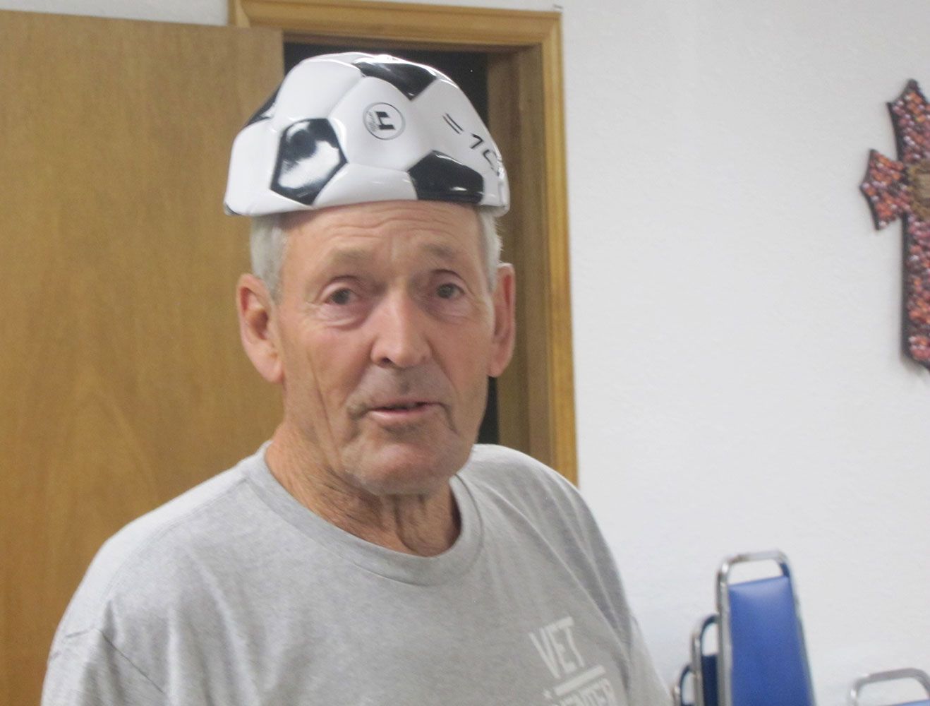 A person wears a soccer ball-patterned cap, standing in a room with a wooden doorway and a cross on the wall.