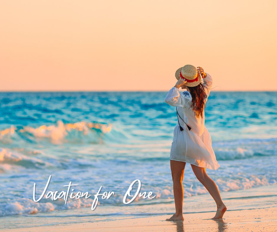 Woman in white dress and straw hat on a beach at sunset, looking out at the ocean.