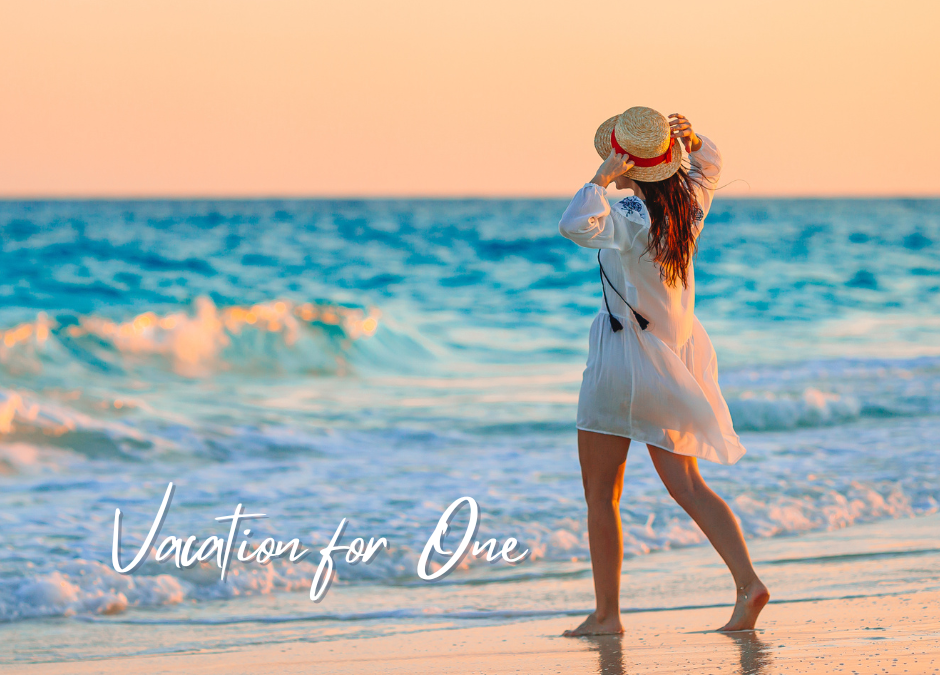 Woman in a white dress and straw hat walks on a beach at sunset. The ocean and sky are colorful.