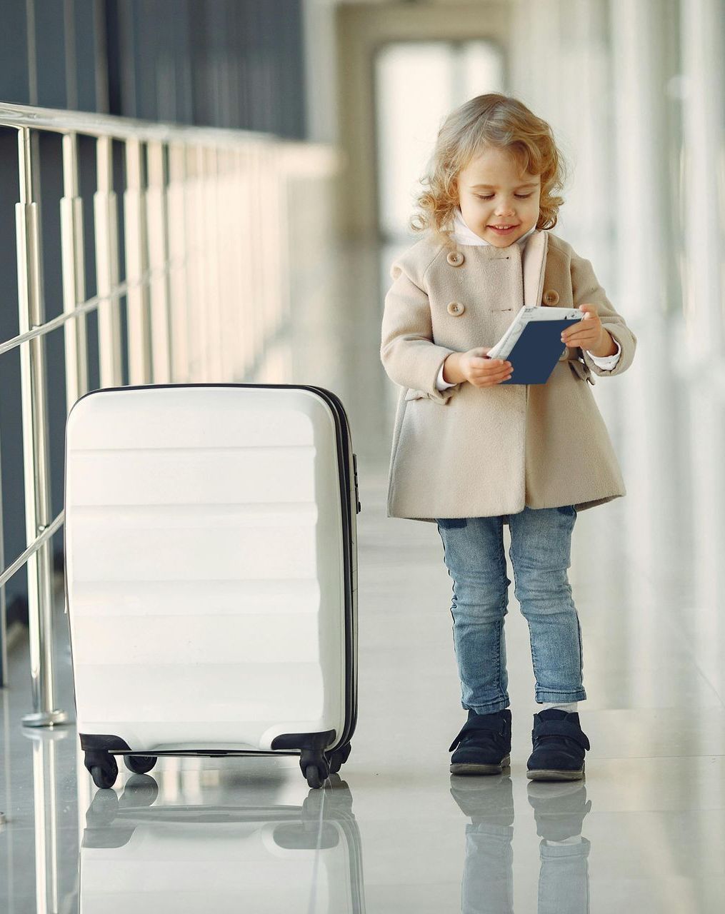 A little girl is standing next to a suitcase and holding a passport.