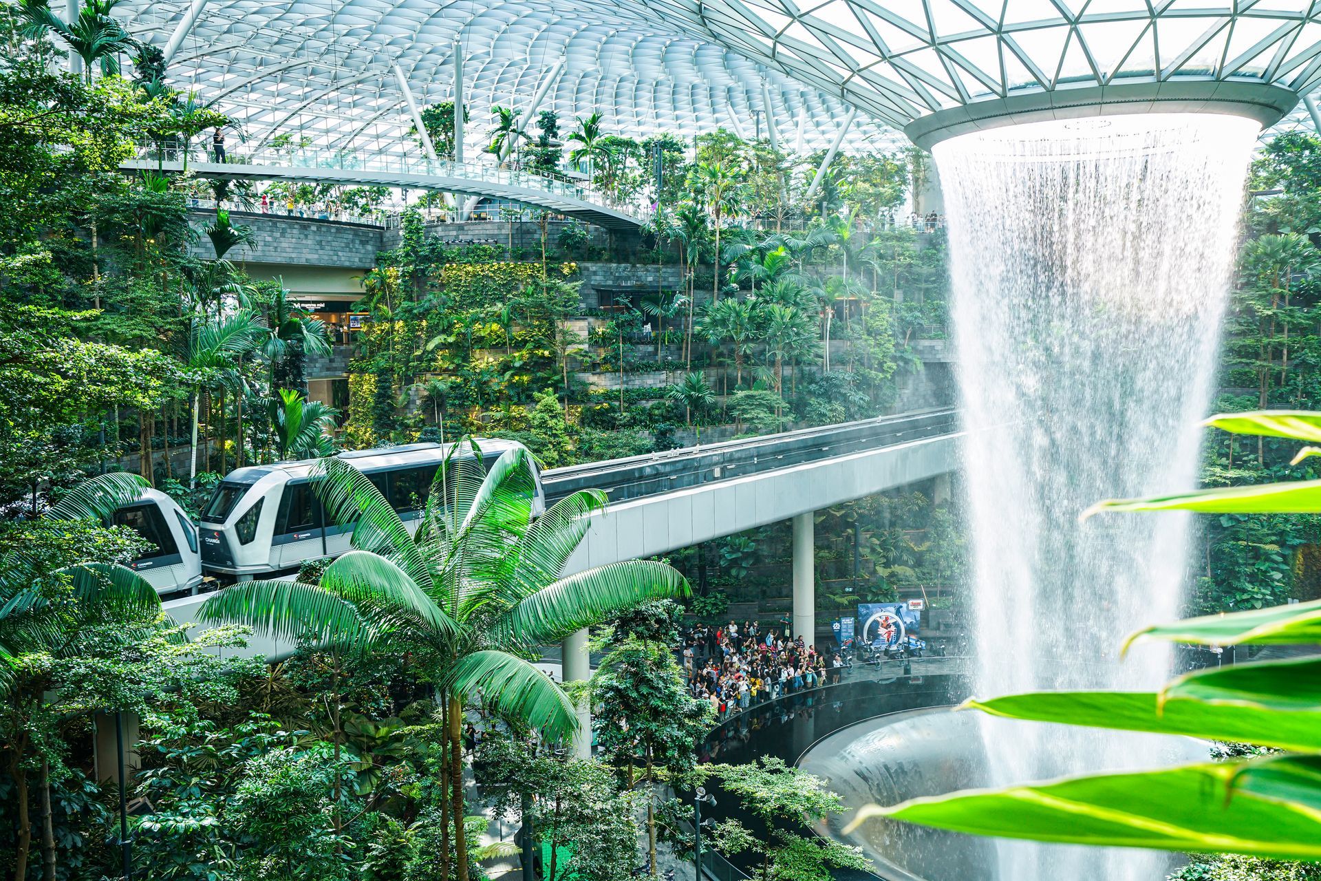 Train traveling past a large indoor waterfall and lush greenery inside the Jewel Changi Airport, Singapore.