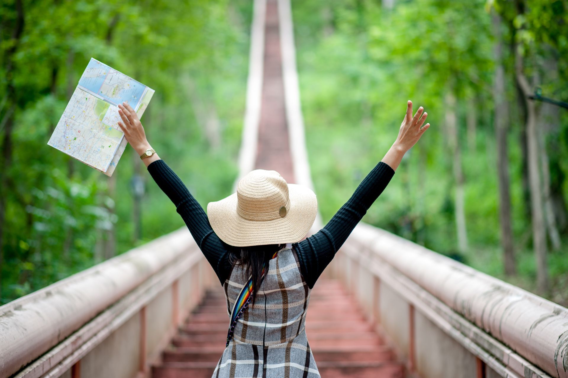 Woman with arms raised, holding a map, stands on a long, red-stepped path through a forest. She wears a hat and plaid top.