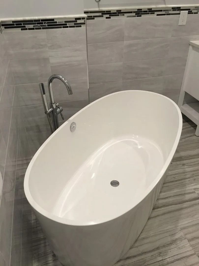 A white, oval freestanding bathtub sits in a bathroom with light gray tiled walls and a dark mosaic accent border.