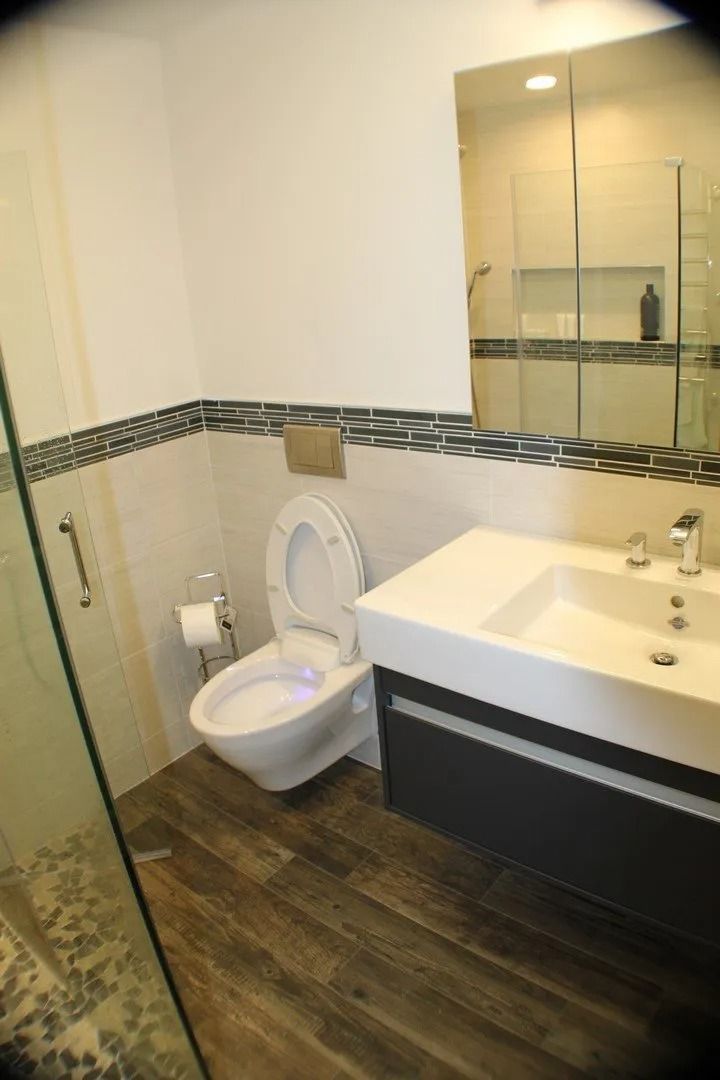 A modern bathroom with a white wall-mounted toilet, dark vanity with a white sink, and wood-look tiled floors.