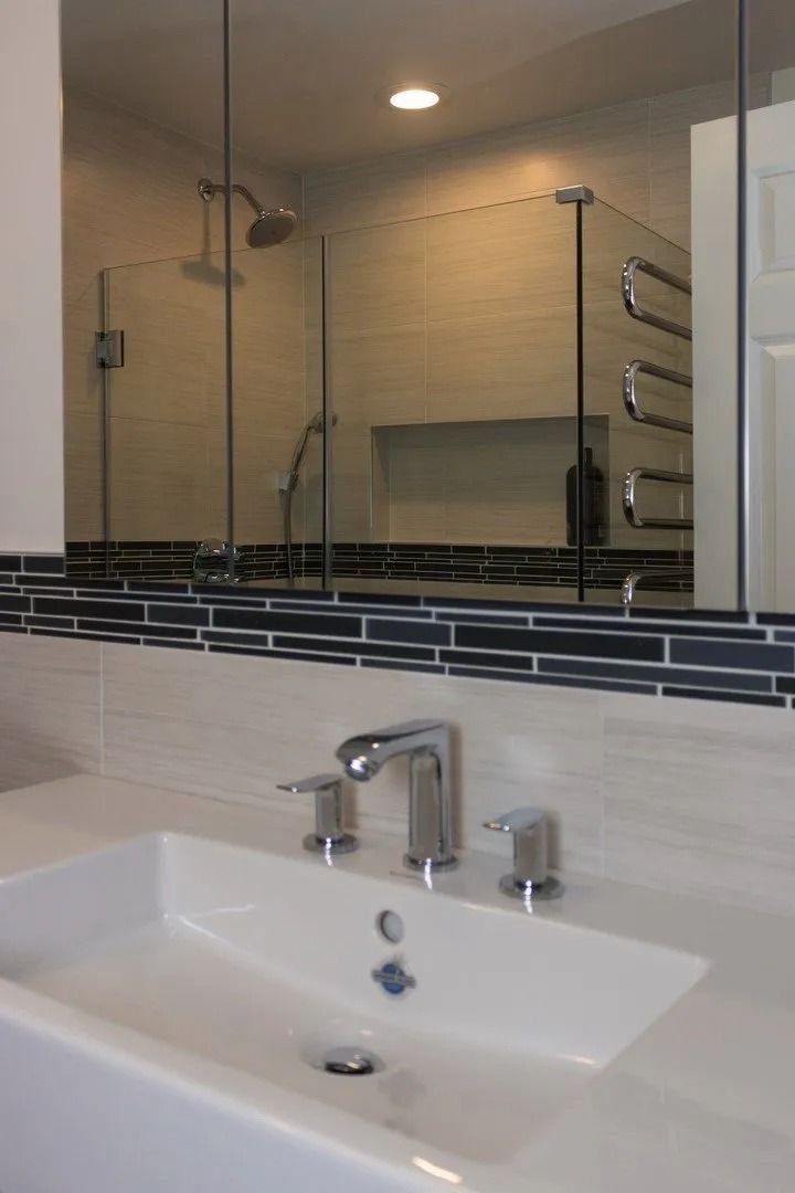 A bathroom sink with a chrome faucet, white basin, and a dark mosaic tile backsplash under a large tri-fold mirror.