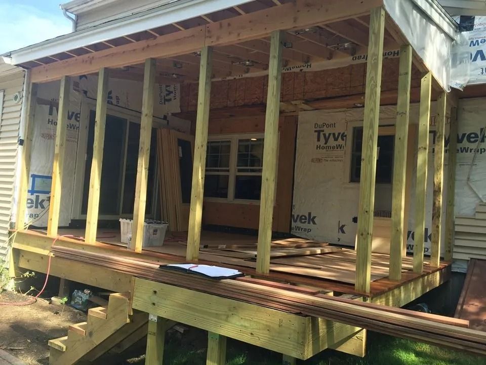 A partially constructed wooden deck and porch frame attached to the side of a house covered in Tyvek wrap.
