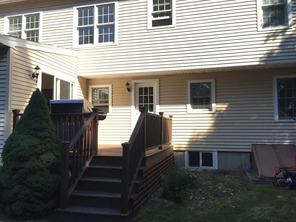 A house exterior with light-colored siding, a wooden deck with stairs, a door, several windows, and an evergreen bush.