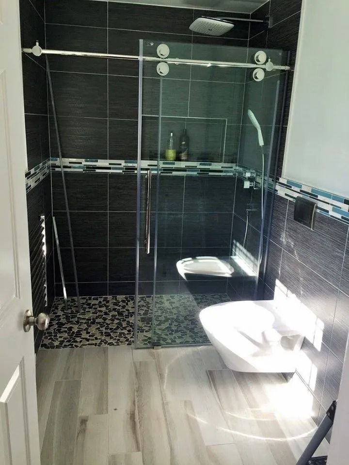 Modern bathroom with gray tiled walk-in shower featuring a pebble floor, glass door, and white wall-mounted toilet.