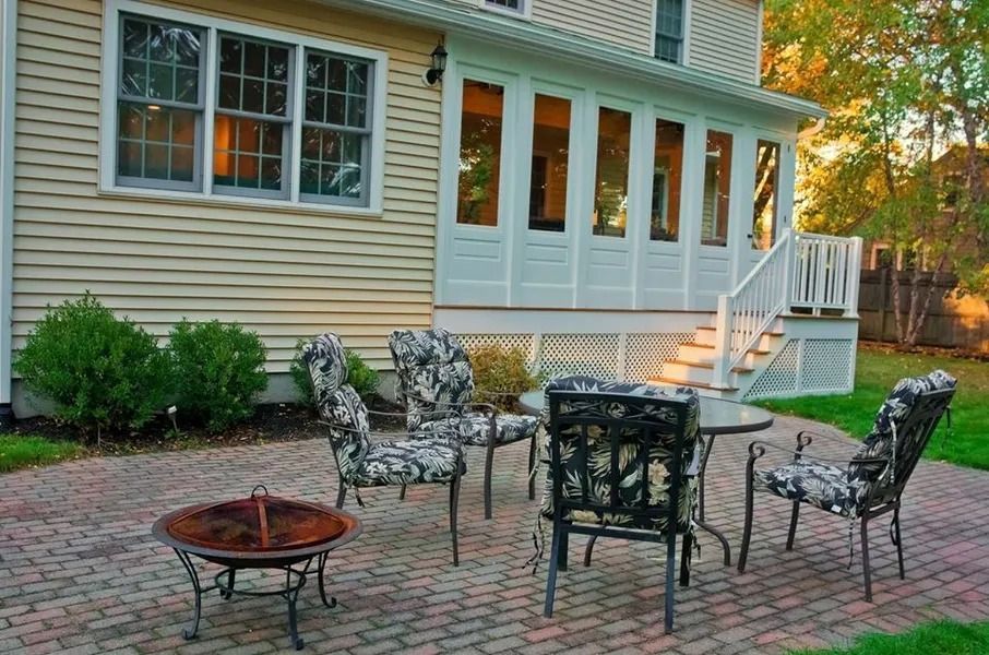 A red brick patio with a metal fire pit and a table set with four chairs outside a yellow house with a white sunroom.