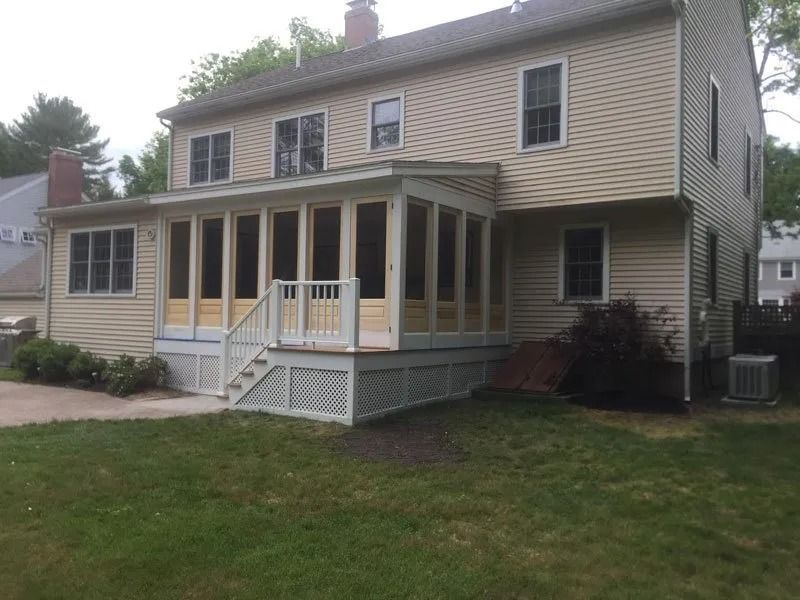 A two-story tan house with a new light-colored sunroom addition, wooden deck, and lattice skirting, set on a grassy lawn.
