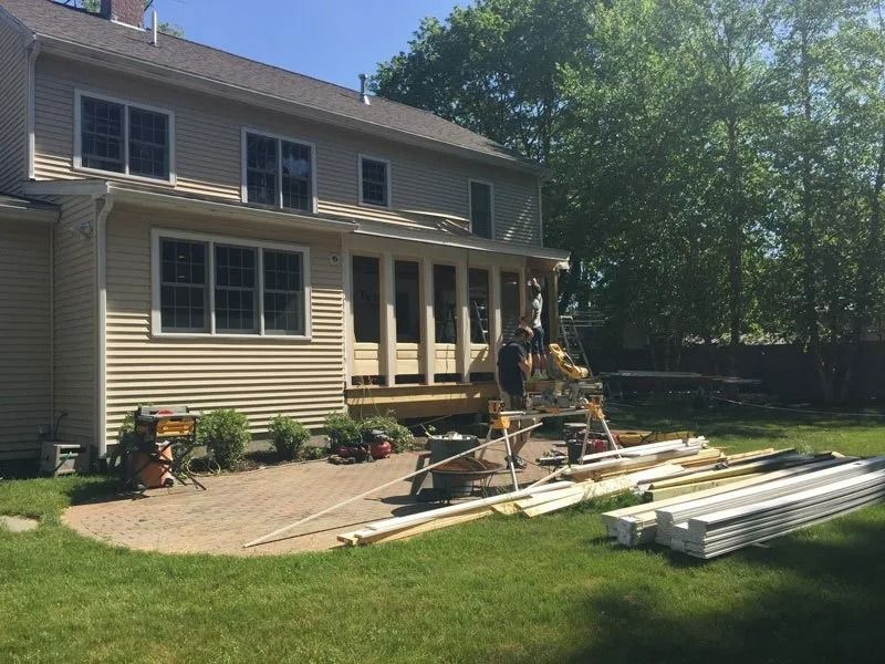 Workers build a wooden deck onto the back of a beige two-story house with a brick patio and lawn.