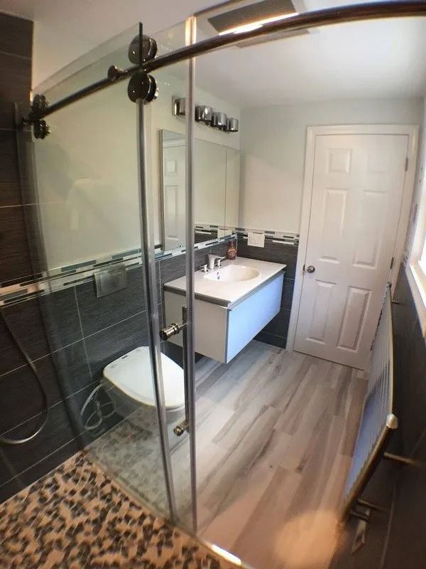 A modern bathroom with a glass-enclosed shower, a floating white vanity, light wood-look floors, and dark tiled walls.
