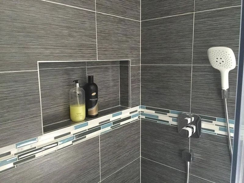 A shower stall with dark gray textured tiles, a recessed shelf containing two bottles, and a white handheld showerhead.