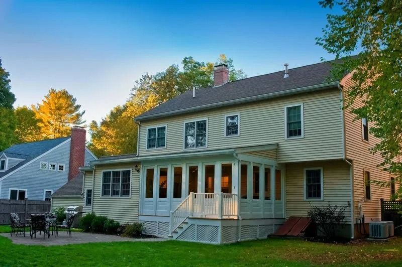 A two-story beige house with a glass-enclosed sunroom and a stone patio on a grassy lawn during autumn.