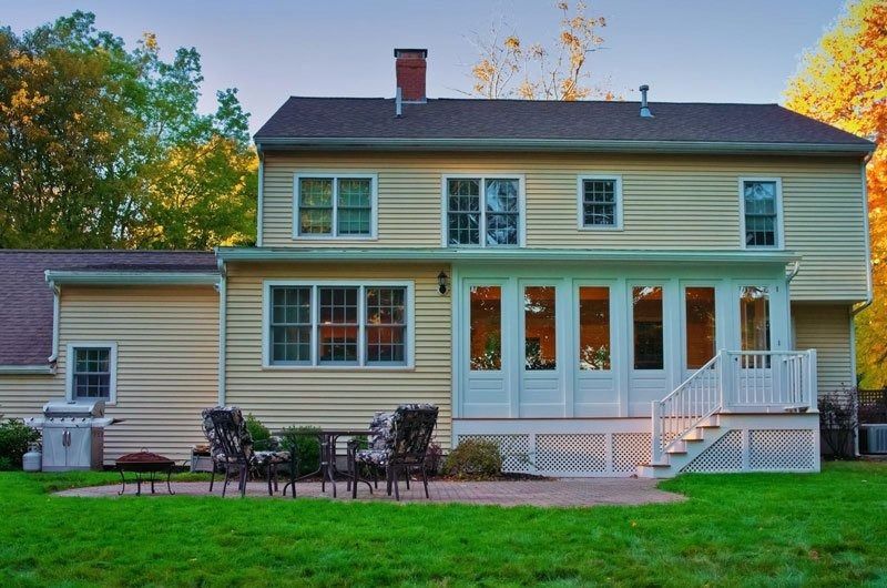 A tan two-story house with a white sunroom addition, a back patio with chairs, and a grassy yard in autumn.