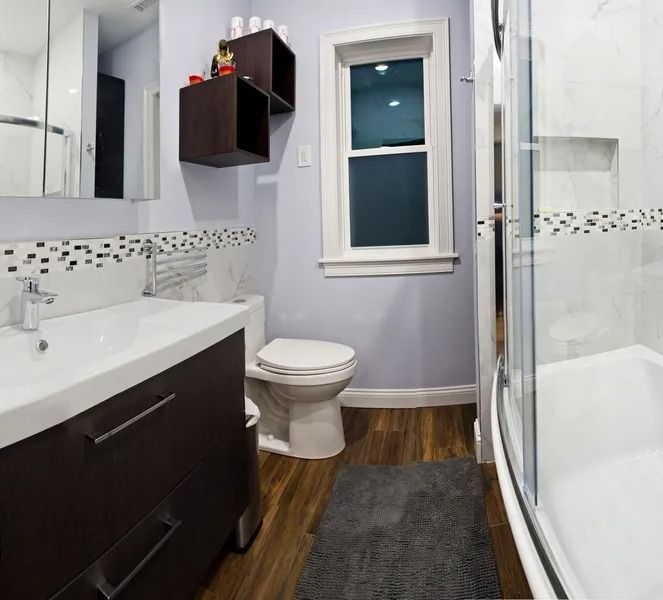 A modern bathroom featuring a dark wood vanity, white toilet, glass shower, and wood-look flooring under a window.