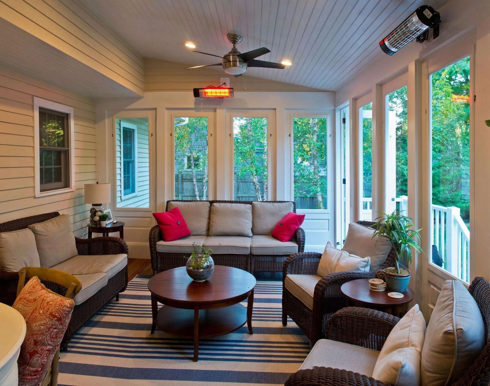 A cozy, light-filled sunroom featuring wicker furniture, a round coffee table, and a striped rug under a ceiling fan.