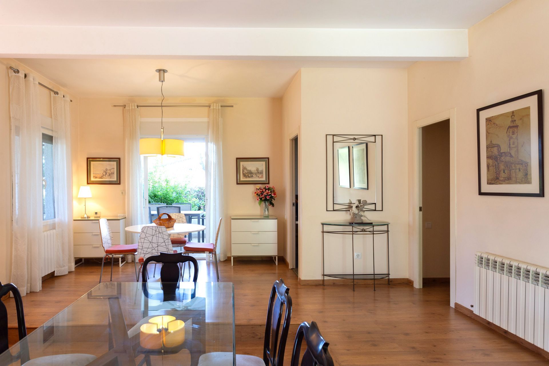 Bright dining room with a glass table, wooden floors, and sunlight from a back doorway.