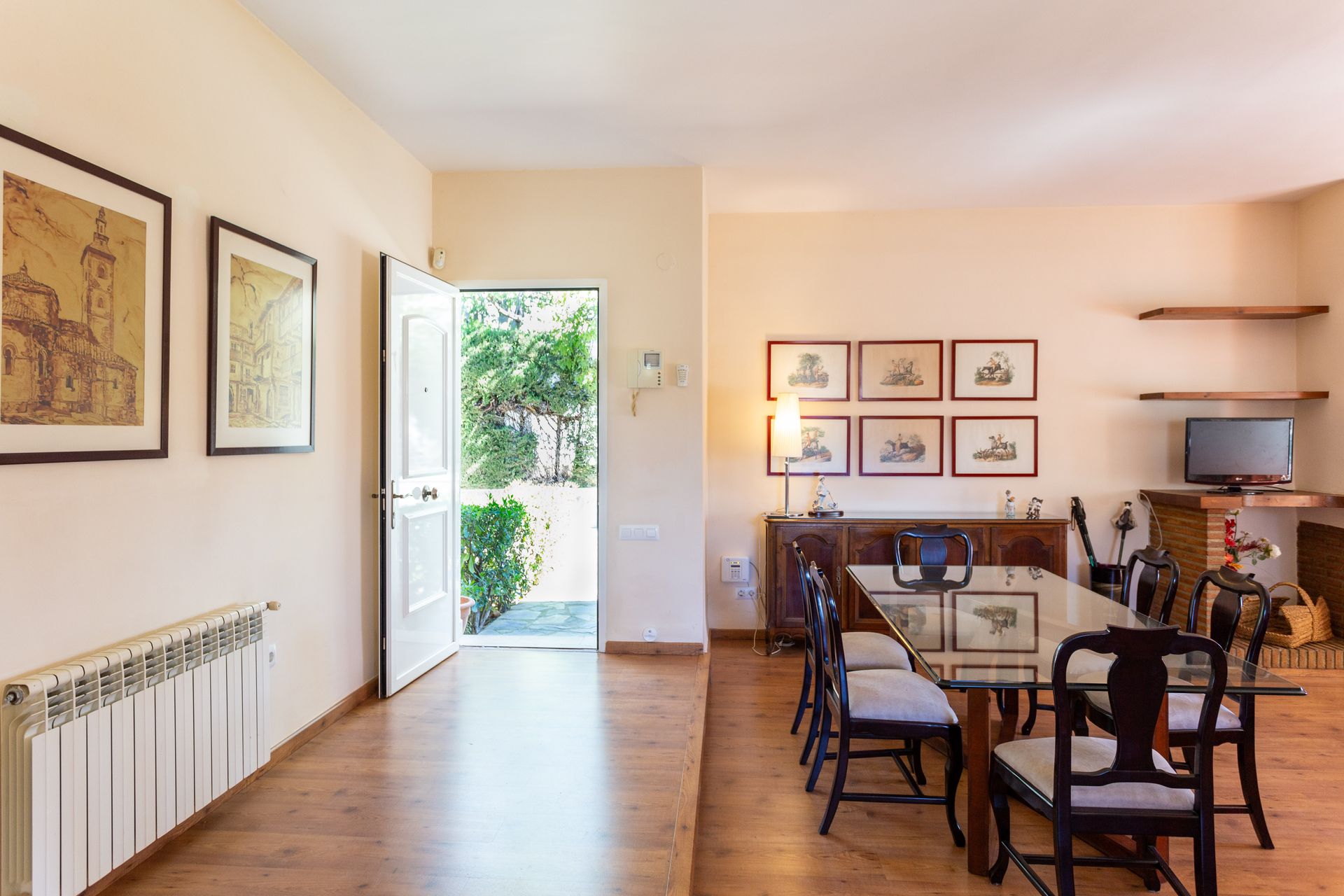 Bright dining room with wooden table, chairs, framed art, and an open door to a sunny garden.