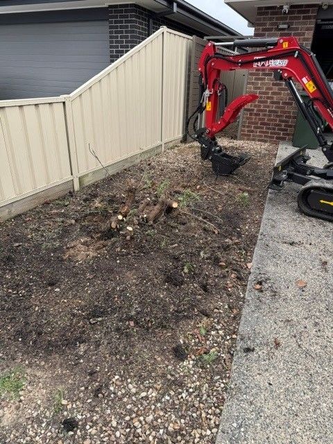 Mini excavator digging out a garden bed beside a house and fence. Preparing for some stone