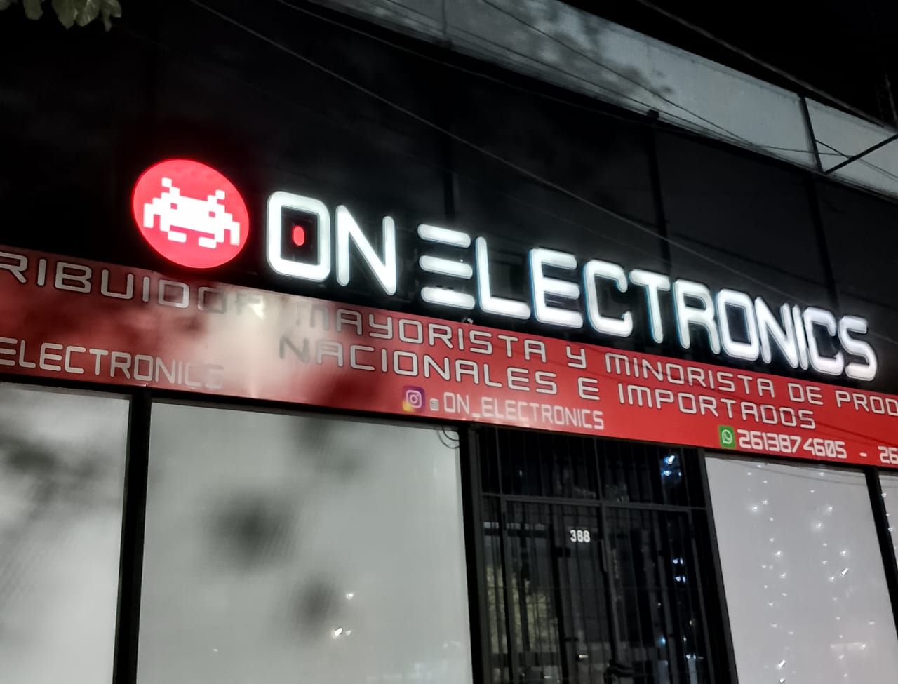 Tienda ON ELECTRONICS con el logo rojo y blanco de una criatura pixelada. Edificio negro de noche.