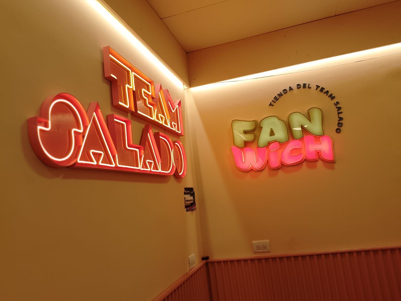 Letreros de neón: “TEAM CALAD”, “FAN WICH” y “tienda del team salado” en una pared de color amarillo claro.
