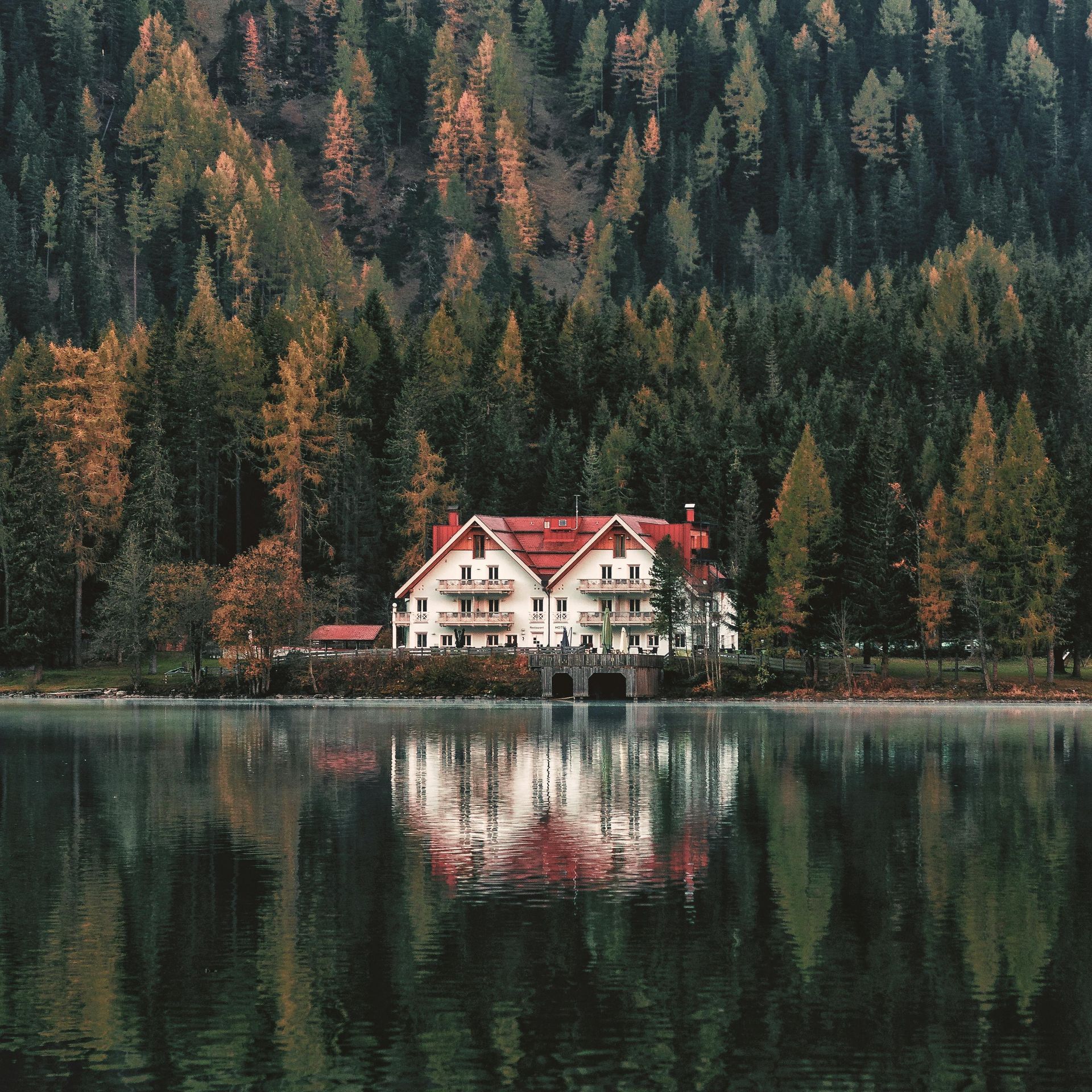 Lakeside house with red roof, reflecting in calm water, surrounded by forest with fall colors.