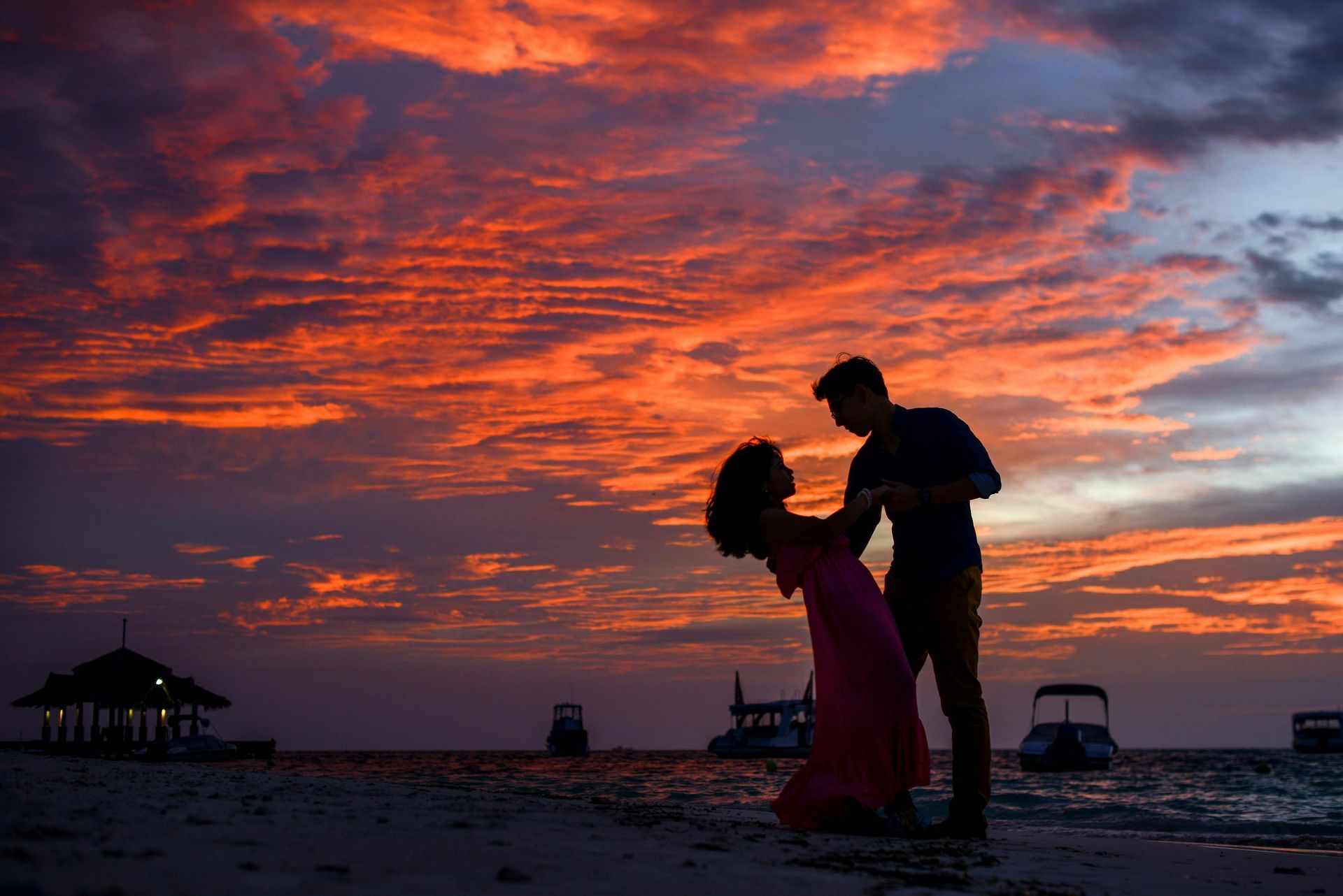 Couple dancing on beach at sunset, silhouetted against vibrant orange and purple sky. Boats in the water.