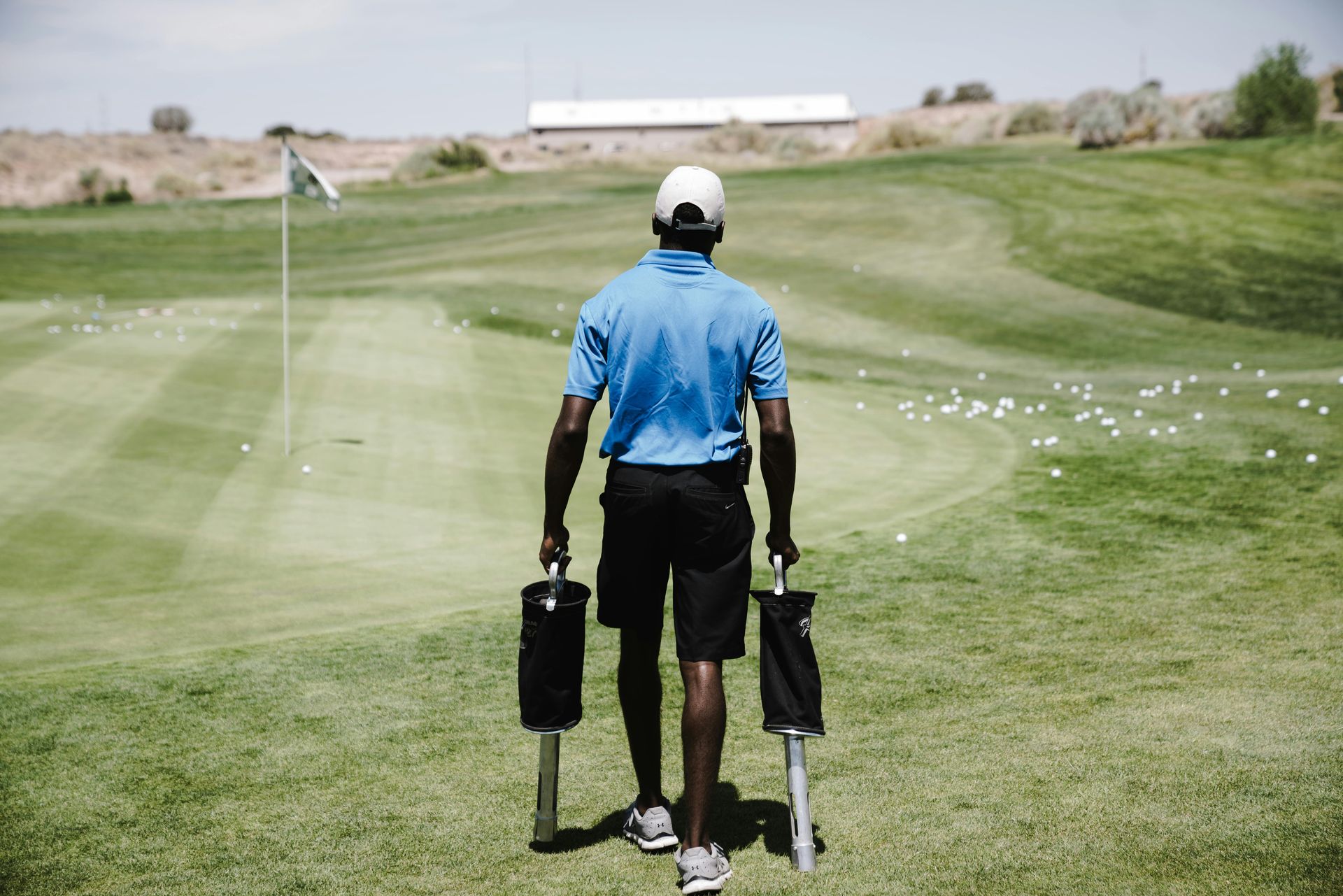 Person walking on a golf course, carrying two black golf club bags, towards a flag.