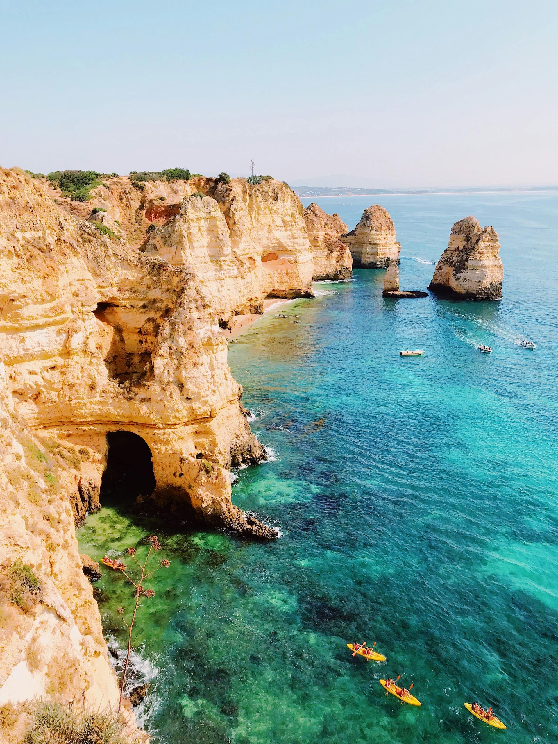 Cliffs overlook turquoise ocean, with kayaks dotting the water, caves visible in the rock face, and a clear blue sky.