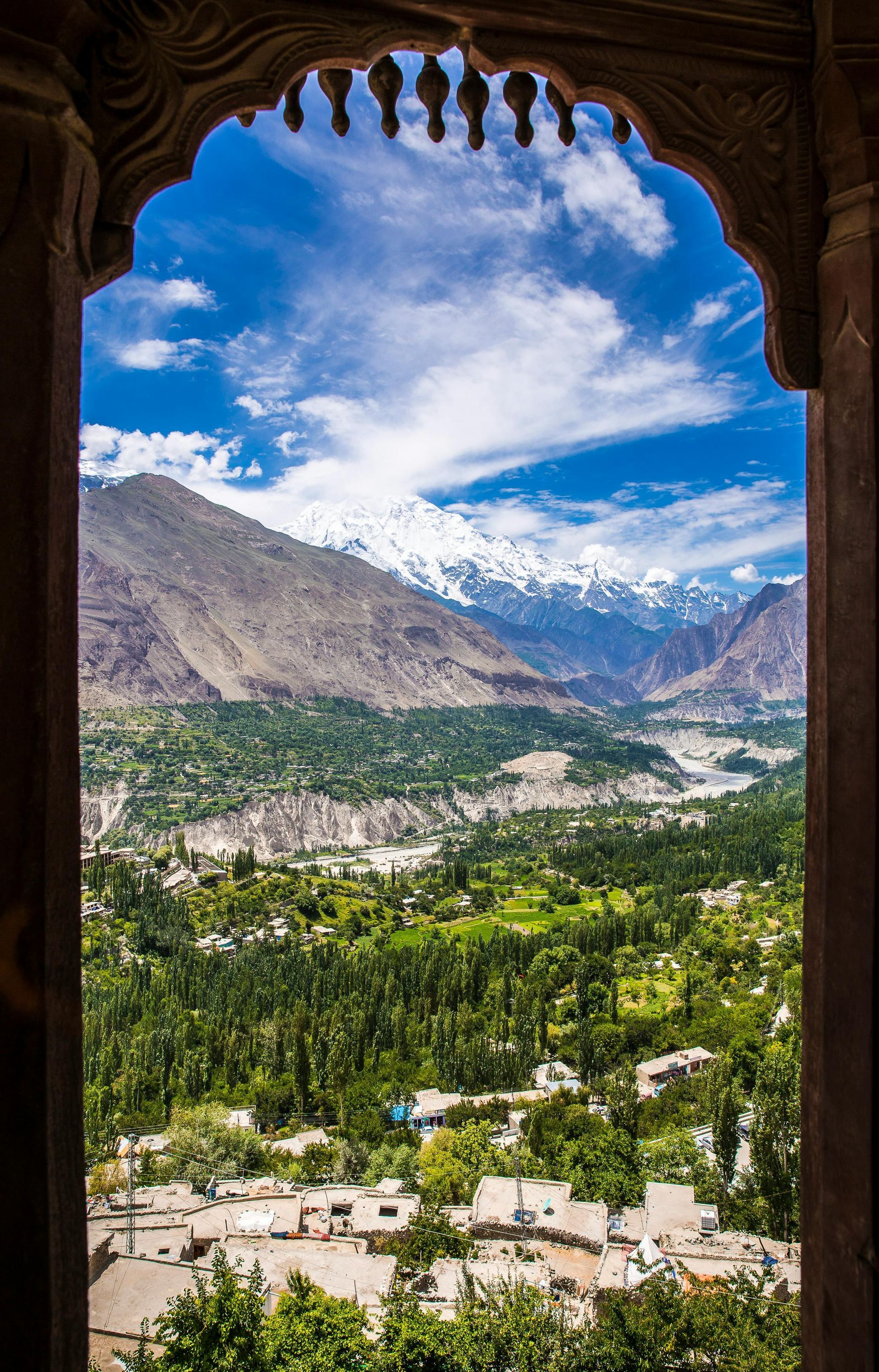 Valley and snow-capped mountains viewed through an ornate archway. Green trees, white rooftops, and blue sky.