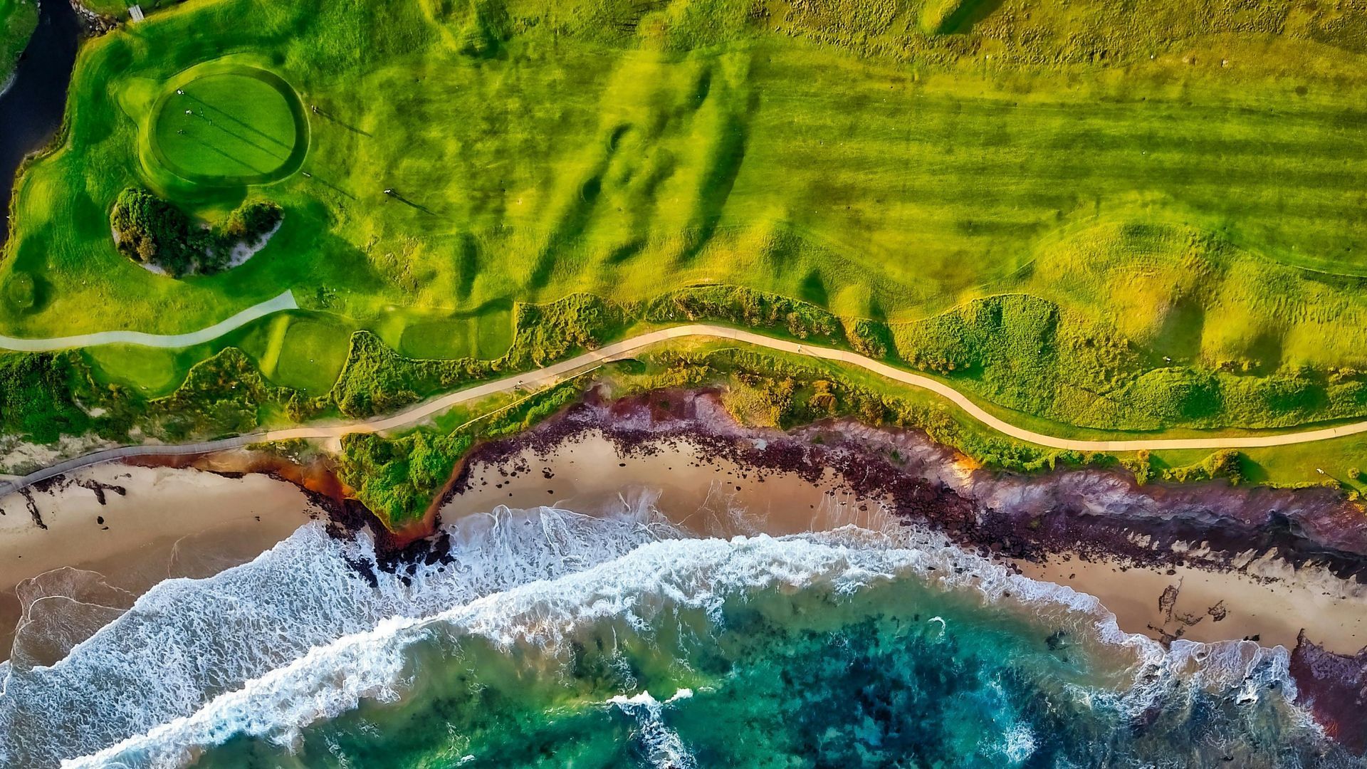 Aerial view of a green grassy cliff, sandy beach, and turquoise ocean with waves.