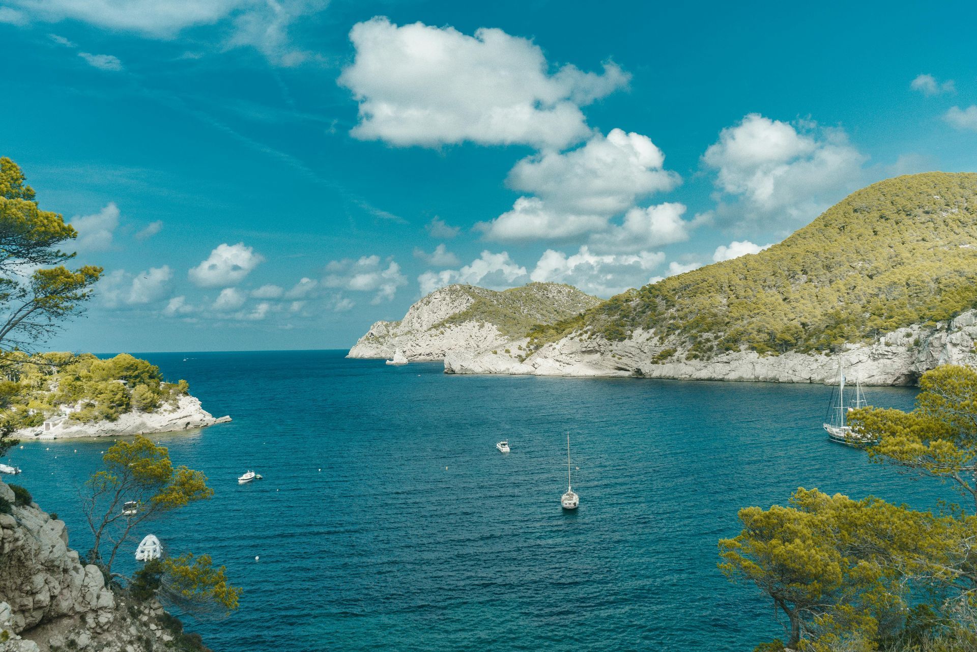 Bay with turquoise water, boats, and green hills under a blue sky with white clouds.