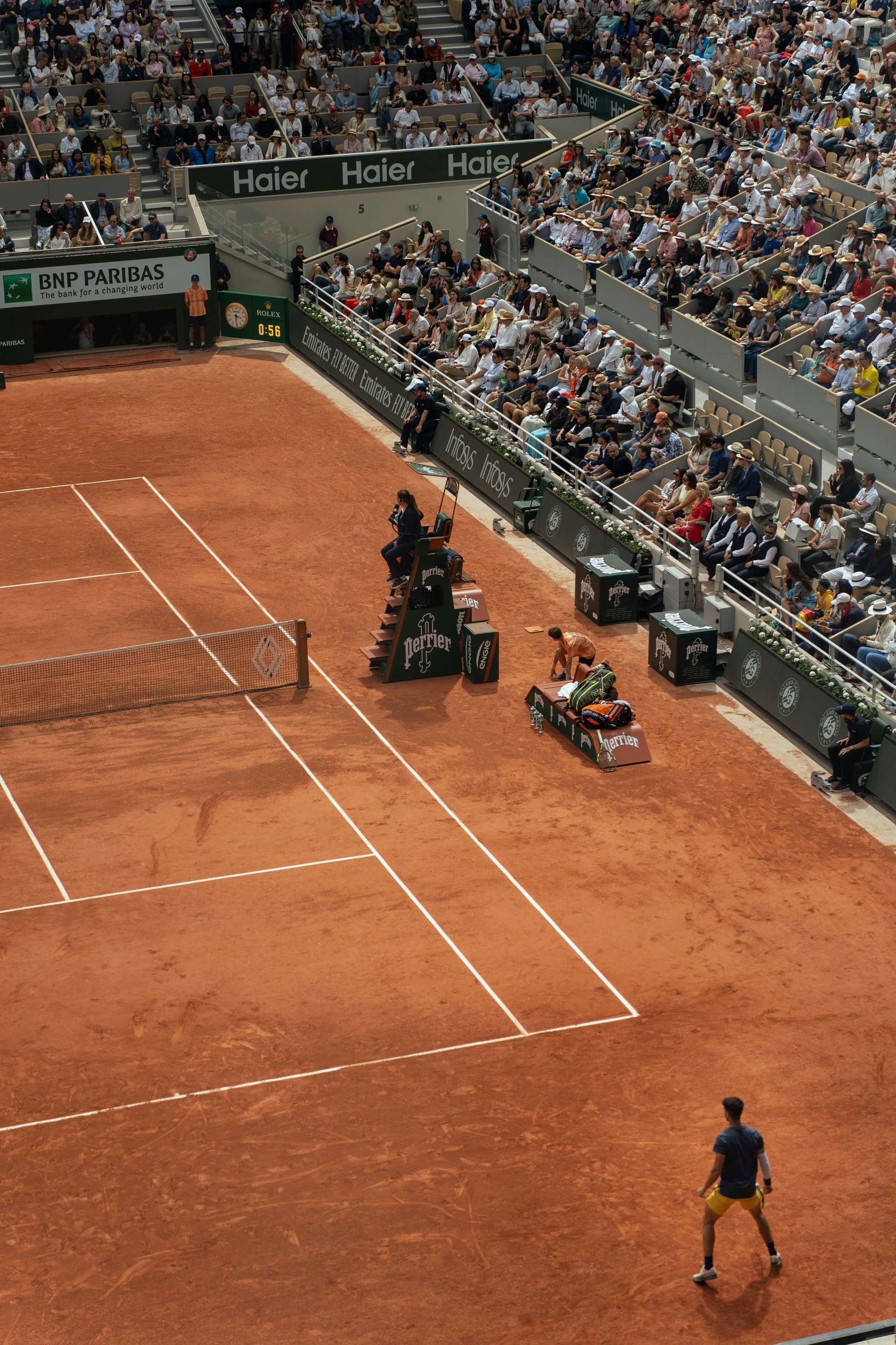 Tennis player on a red clay court, ball in play, crowd in the background, official with a machine.