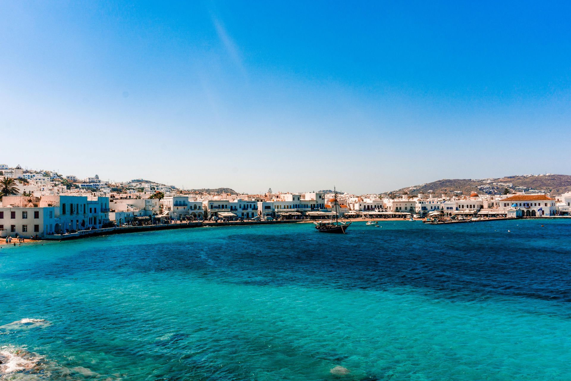 Bright turquoise water, white buildings line the coast under a clear blue sky in Mykonos, Greece.