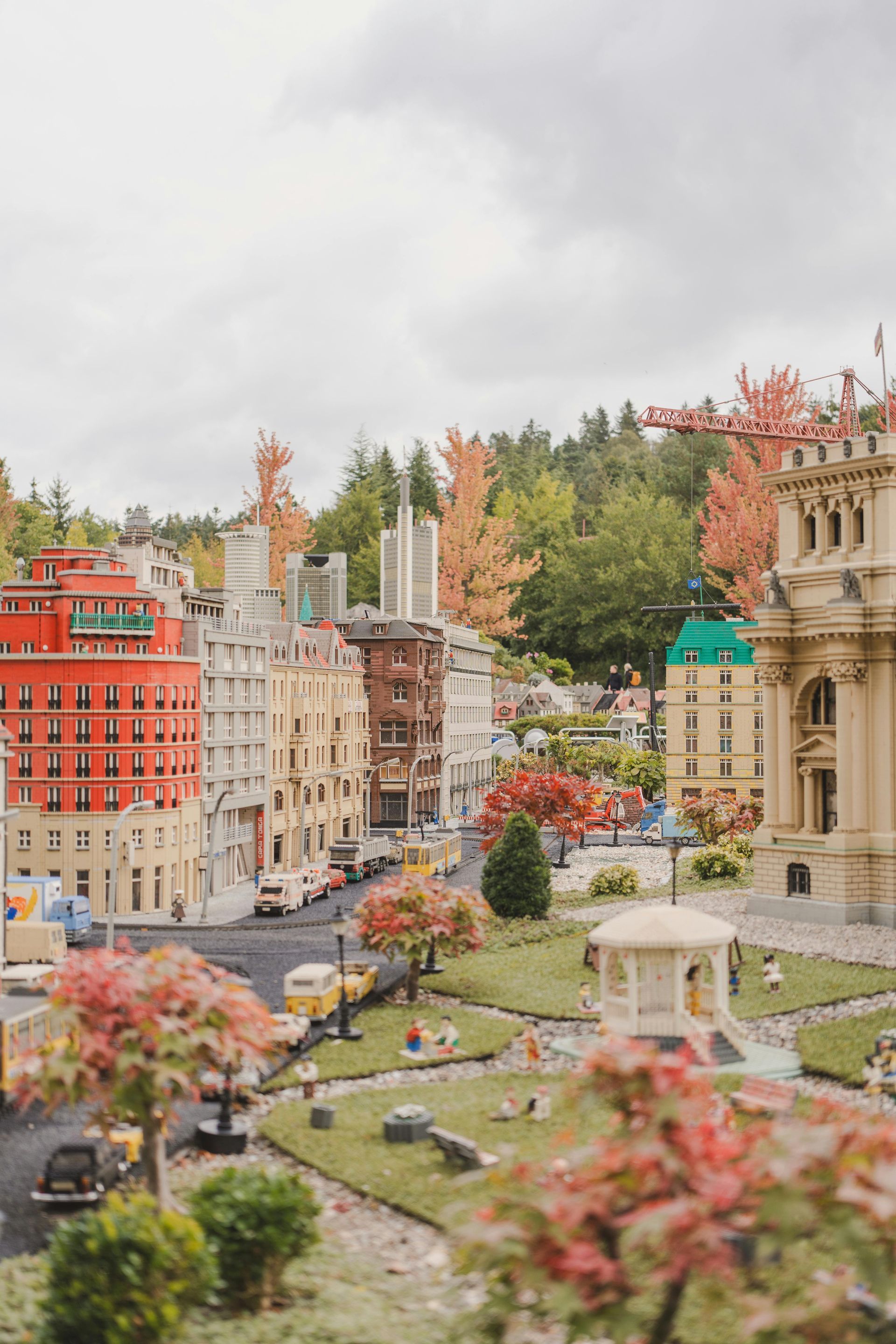 Lego city with buildings, trees, and cars. Autumn foliage and a cloudy sky are in the background.
