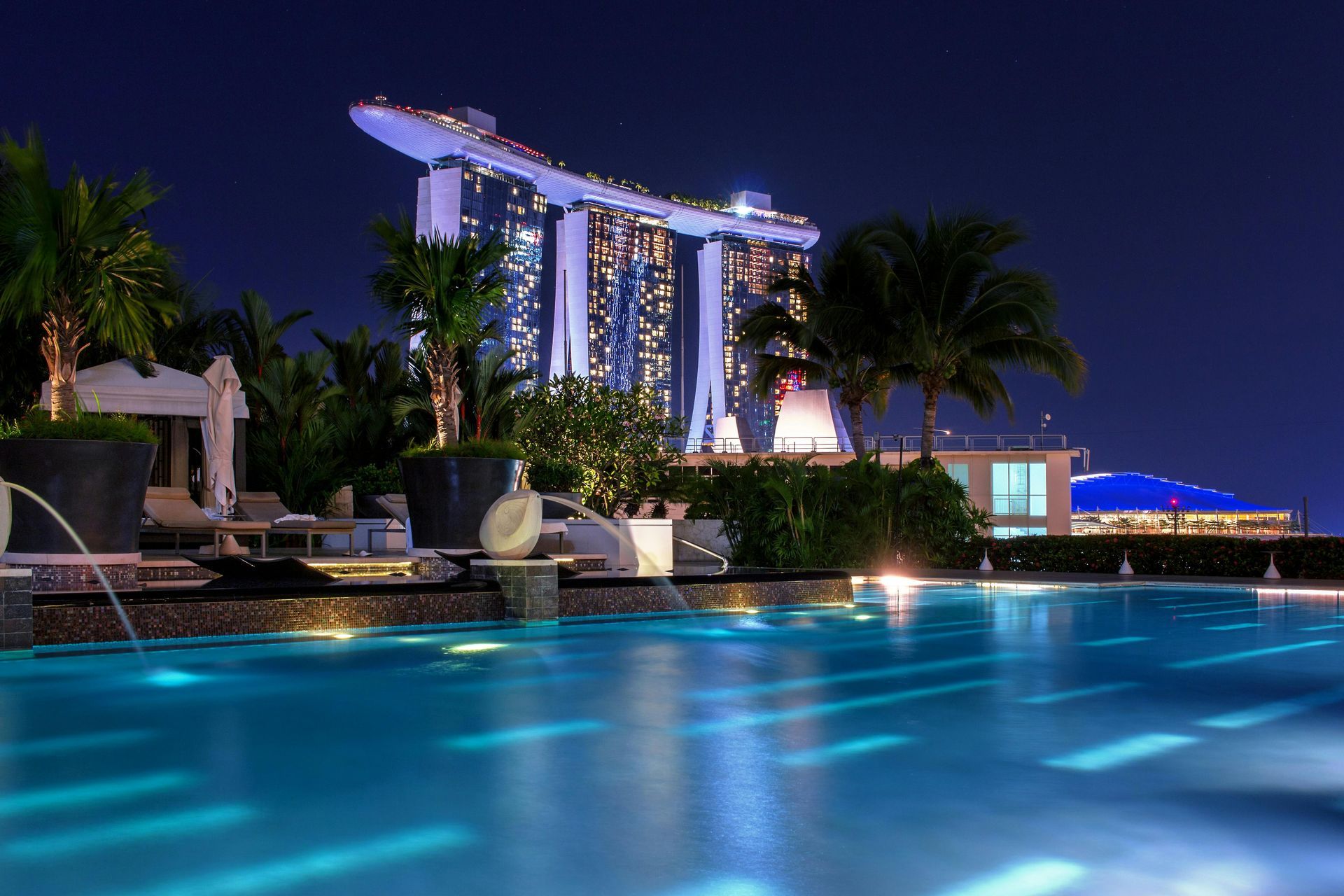 Night view of a rooftop pool overlooking the illuminated Marina Bay Sands hotel in Singapore. Blue water and sky.