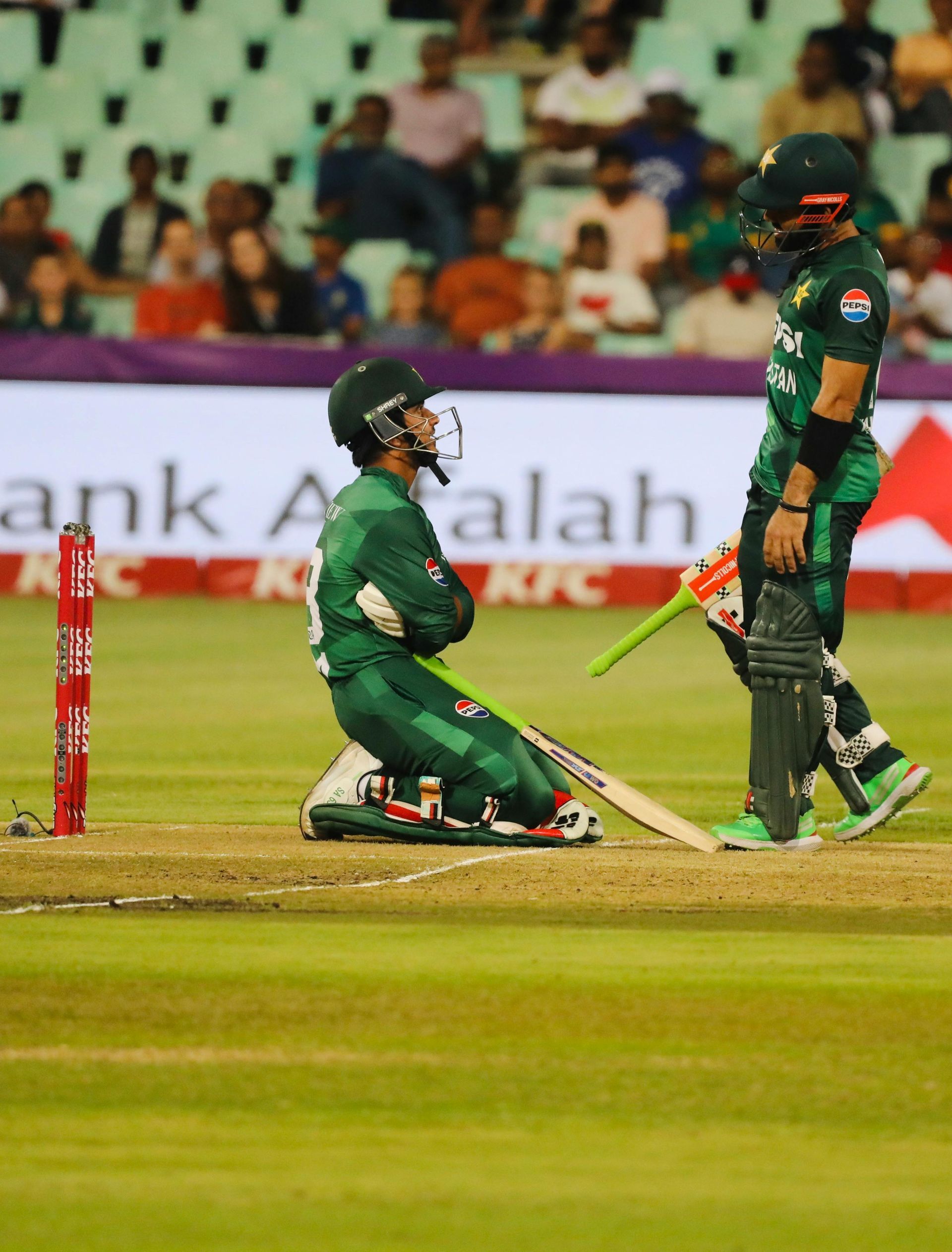 Cricket players, one kneeling, the other standing, on a green field. Players wearing green uniforms, stadium in background.